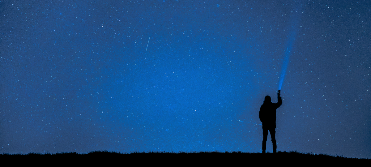 A silhouette of a person standing on a hill against a vast, starry night sky. The person is holding up a flashlight, projecting a powerful blue beam of light into the dark expanse.