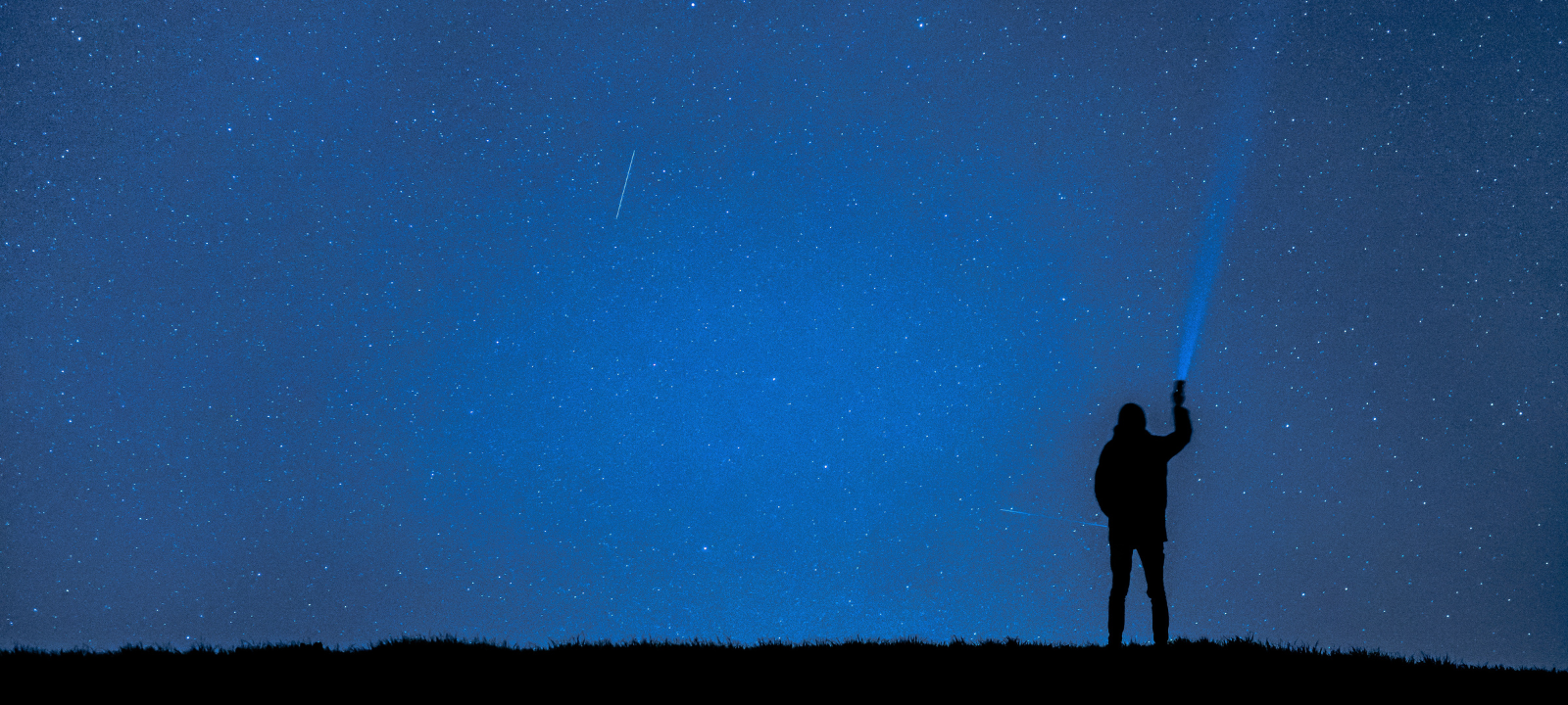 A silhouette of a person standing on a grassy hill under a starry night sky. The person is holding up a flashlight, projecting a blue beam of light into the dark, celestial expanse.
