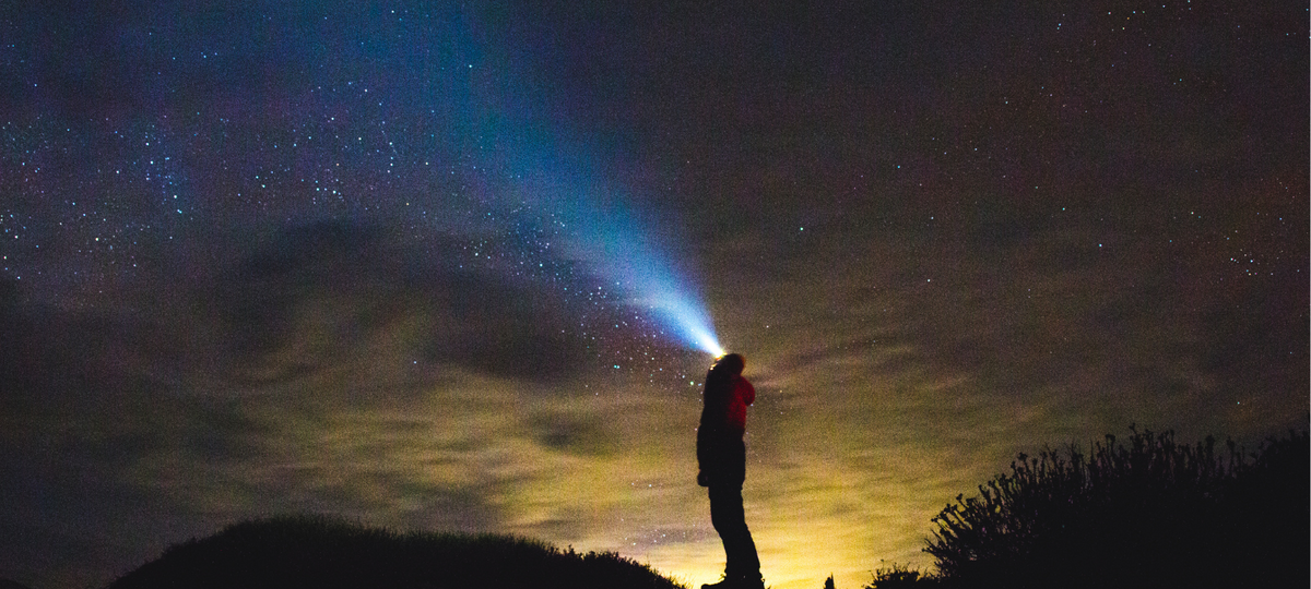 A silhouette of a person standing on a dark ridge, using the flashlight to shine a bright beam of light into a starry night sky. 